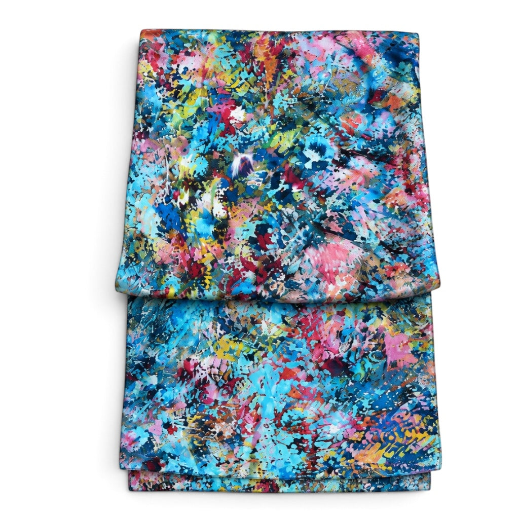 A folded textile item, likely a blanket or throw, featuring a vibrant abstract watercolor paint splatter pattern in shades of blue, pink, red, yellow, and green on a white background.