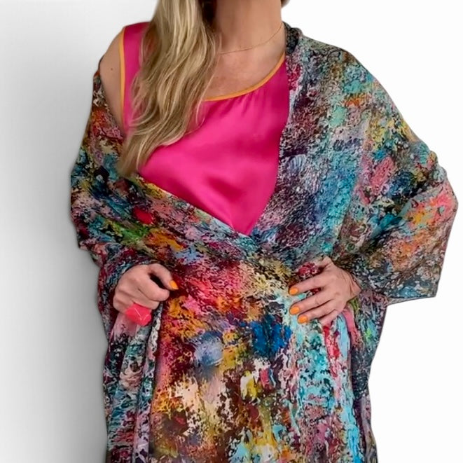 A woman wearing a vibrant, multicolored abstract print shawl or wrap over a hot pink satin top, shown from the neck down against a plain white background.