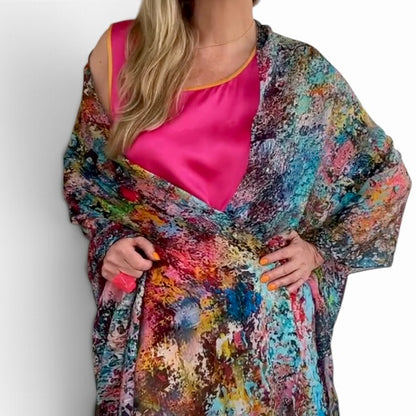 A woman wearing a vibrant, multicolored abstract print shawl or wrap over a hot pink satin top, shown from the neck down against a plain white background.