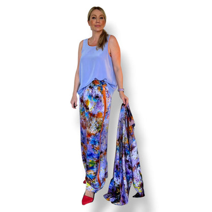 A woman modeling a light blue sleeveless top and vibrant multicolor abstract print pants, holding up a matching multicolor fabric scarf or shawl against a plain white background.
