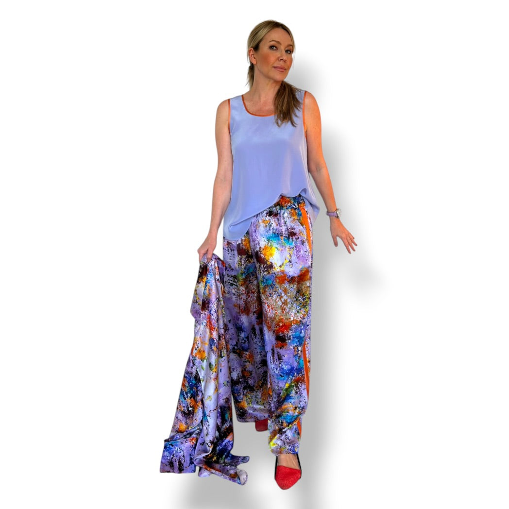 A woman modeling a sleeveless light blue top and wide-leg draped pants with a colorful abstract print, paired with red shoes, against a plain white background.