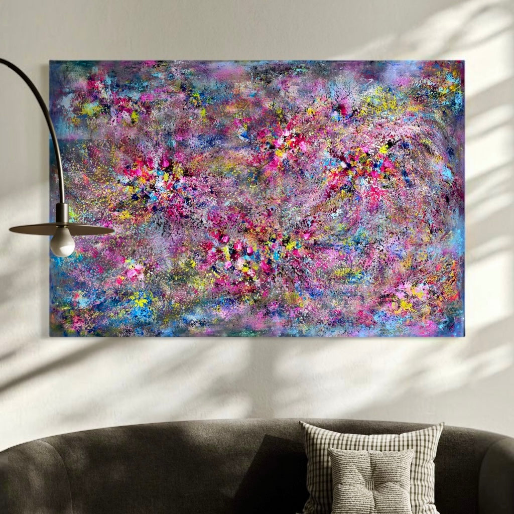 A large rectangular abstract wall art piece with a dense, expressive mix of pink, purple, blue, and yellow brushstrokes, displayed above a dark gray sofa in a styled interior setting with a floor lamp and soft window shadows.
