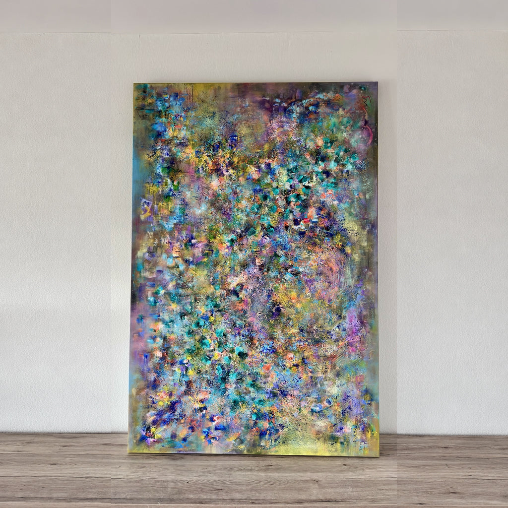 An abstract canvas painting with a dense, textured pattern of blue, green, purple, yellow, and pink brushstrokes, leaning against a white wall on a light wood floor.
