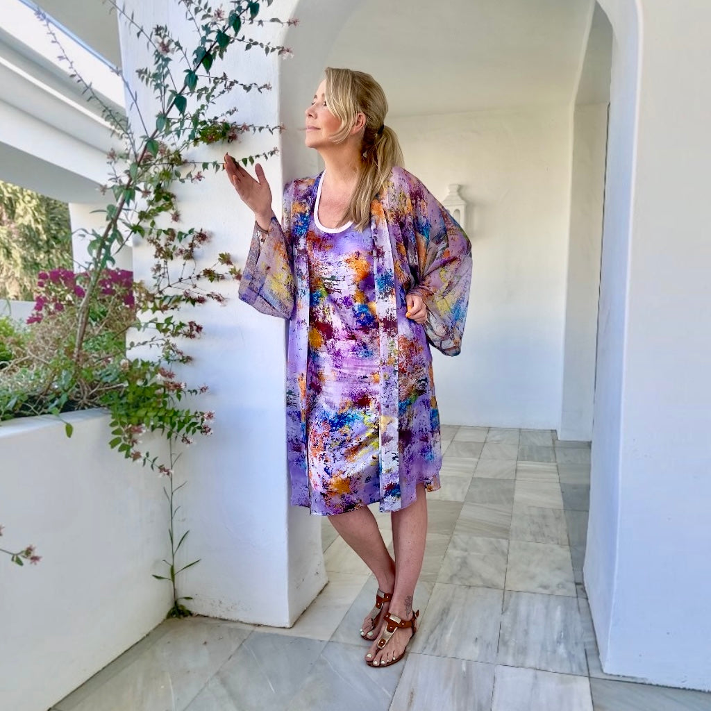 A woman wearing a purple and multicolor abstract print kaftan dress with wide sleeves, standing in a bright, white-walled outdoor corridor with tiled floors and climbing plants.