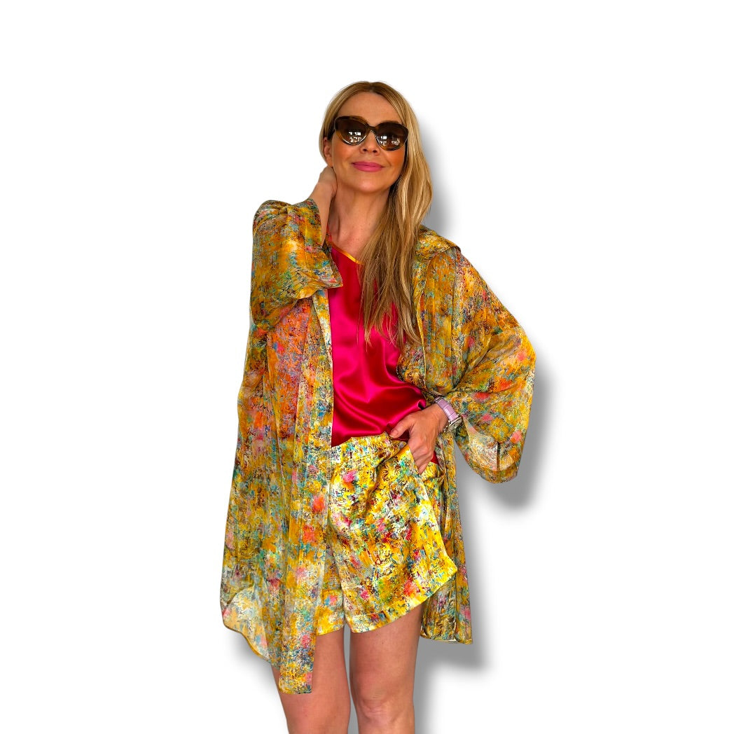 A woman modeling a matching floral kimono jacket and shorts set in yellow, orange, and green, worn over a bright pink satin top and paired with dark sunglasses, against a white background.
