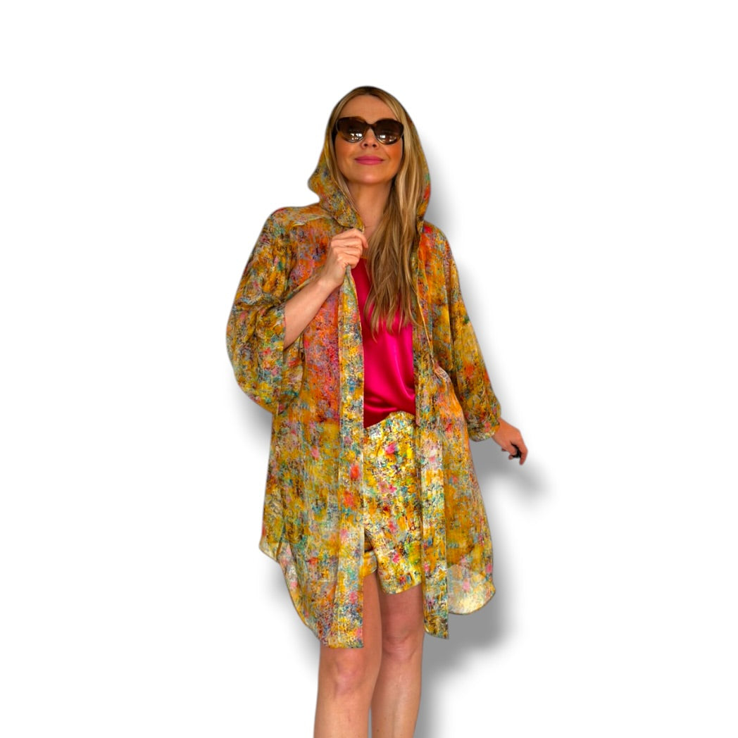 A woman modeling a lightweight hooded kimono-style jacket with a dense multicolor floral print in yellow, orange, green, and blue over a bright pink top, set against a plain white background.