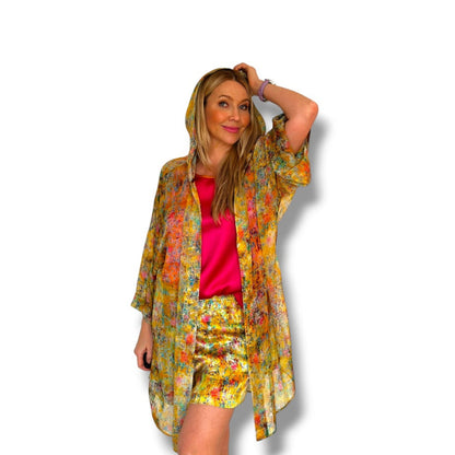 A woman modeling a matching three-piece floral kimono set with a hood, short-sleeve cardigan, and shorts in a vibrant yellow, orange, and red floral print, worn over a hot pink satin top, against a plain white background.