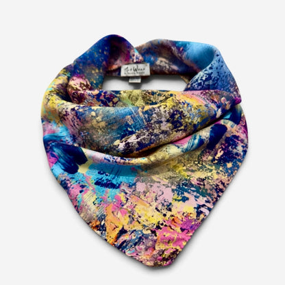 A multicolor abstract painterly print scarf draped in a loop to show its shape and pattern, featuring shades of blue, yellow, pink, and purple on a light gray background.