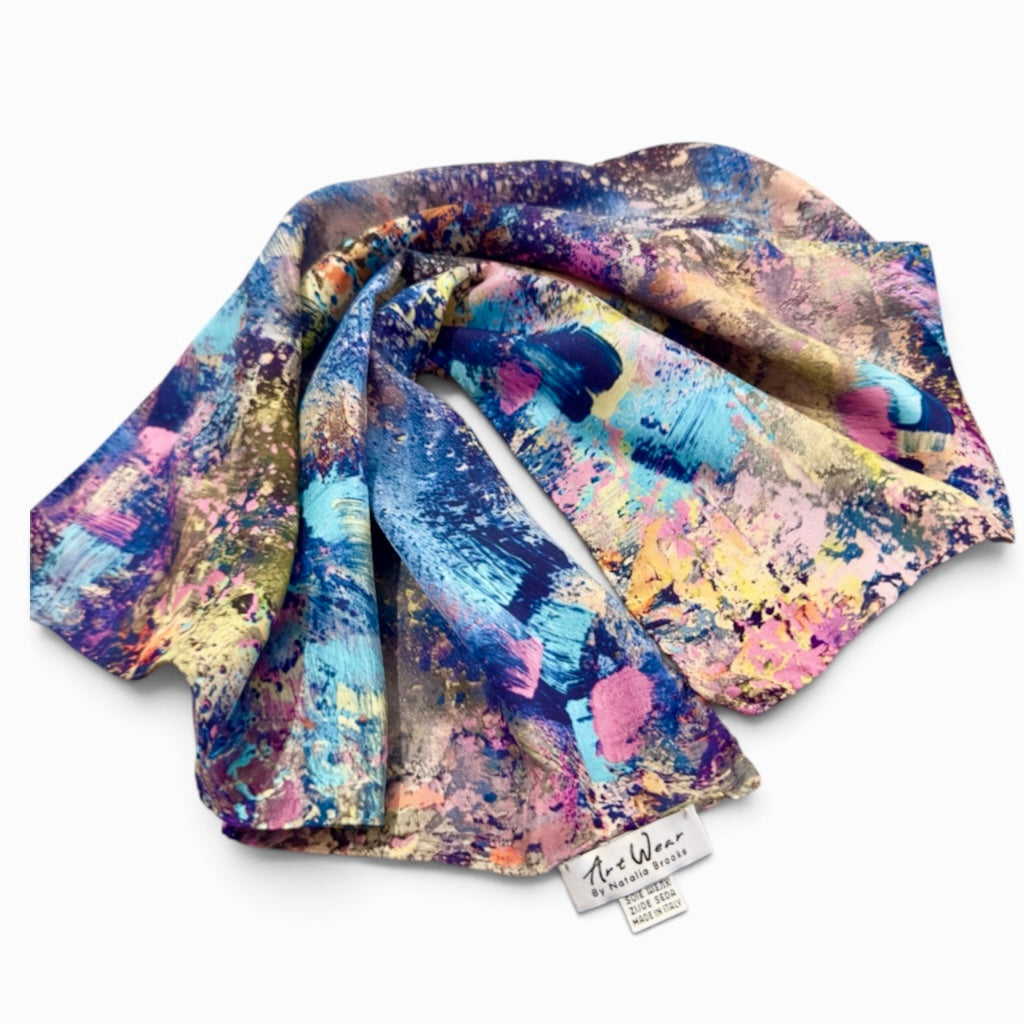 A lightweight scarf with an abstract multicolor paint-splatter pattern, featuring shades of blue, pink, purple, yellow, and beige. A small white fabric tag with the brand name 'The Wear' is visible at the bottom edge, set against a clean white background.