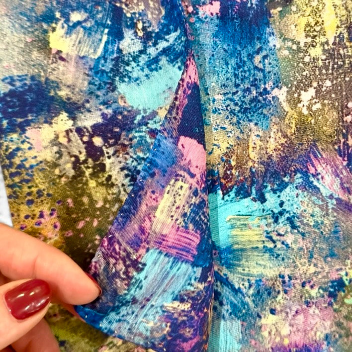 A close-up of a colorful abstract-patterned garment, showing a hand with red painted fingernails holding the fabric to demonstrate its drape and texture.