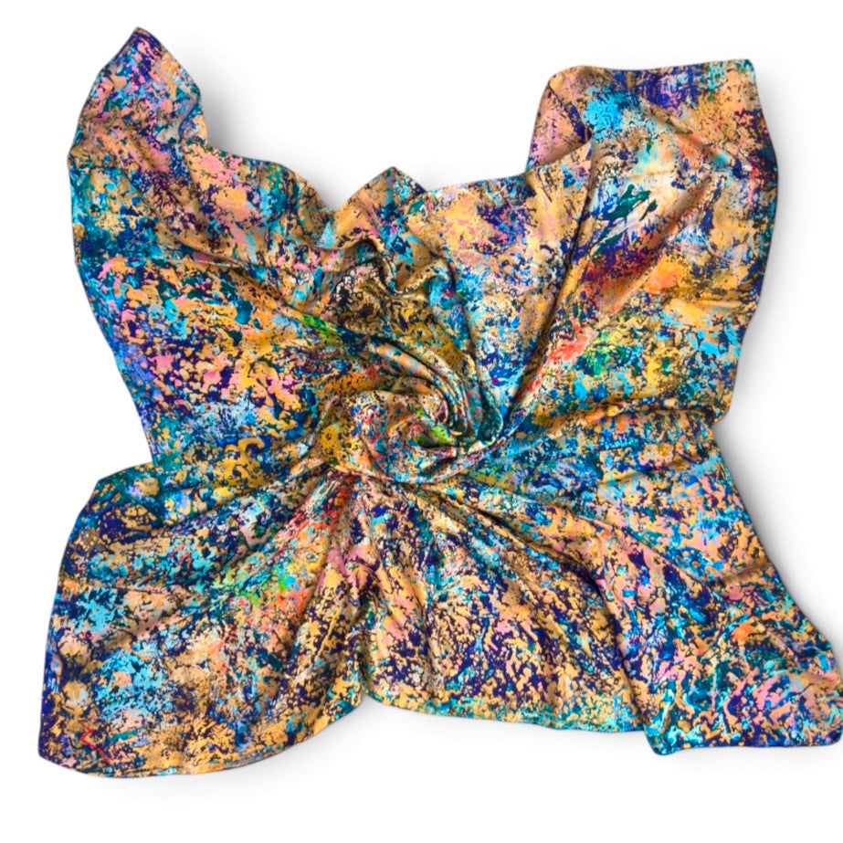 A silk square scarf,  featuring a vibrant multicolor abstract paint-splatter pattern in shades of blue, orange, yellow, pink, and purple, arranged in a large gathered bow on a white background.