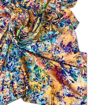 A draped piece of silk fabric with a vibrant, multicolored abstract print featuring splashes of blue, yellow, orange, green, and purple on a white background.
