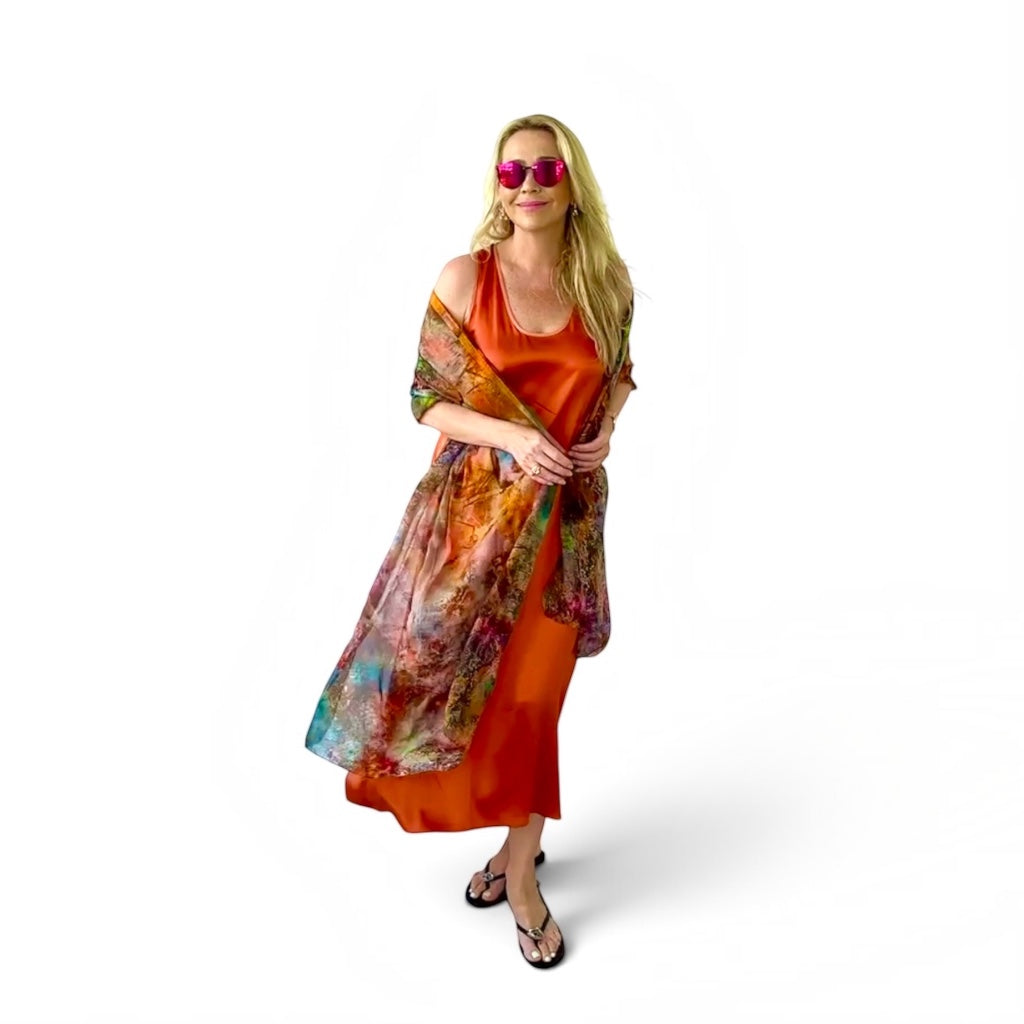 A woman modeling a vibrant orange dress with a colorful, painterly abstract print shawl draped over her shoulders, set against a plain white studio background.