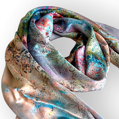 A lightweight silk scarf with a multicolor abstract print, featuring shades of teal, pink, gold, and purple, styled in a loose knot against a light gray background.