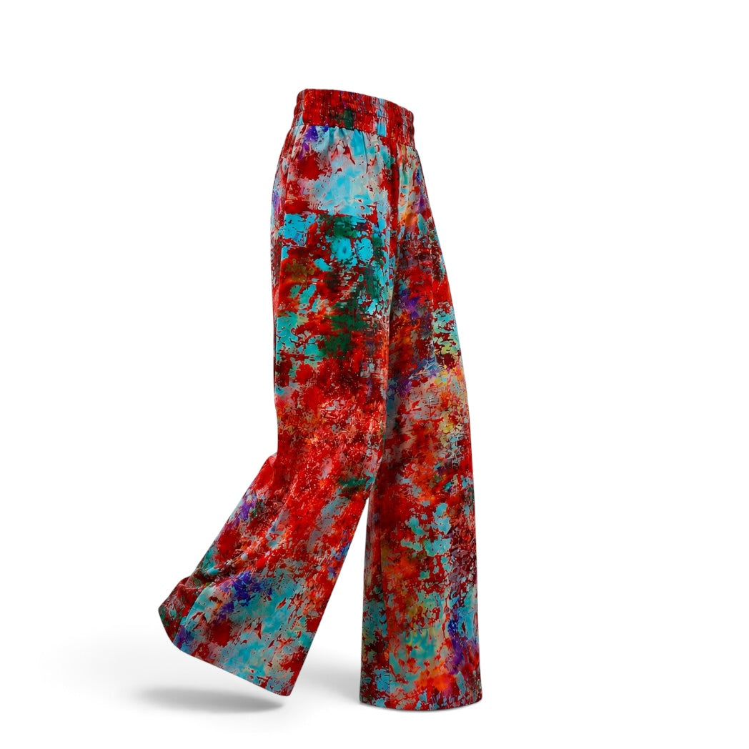 A pair of wide-leg trousers featuring a vibrant, multicolored abstract paint splatter pattern with prominent red, turquoise blue, and orange tones, shown on a clean white background.