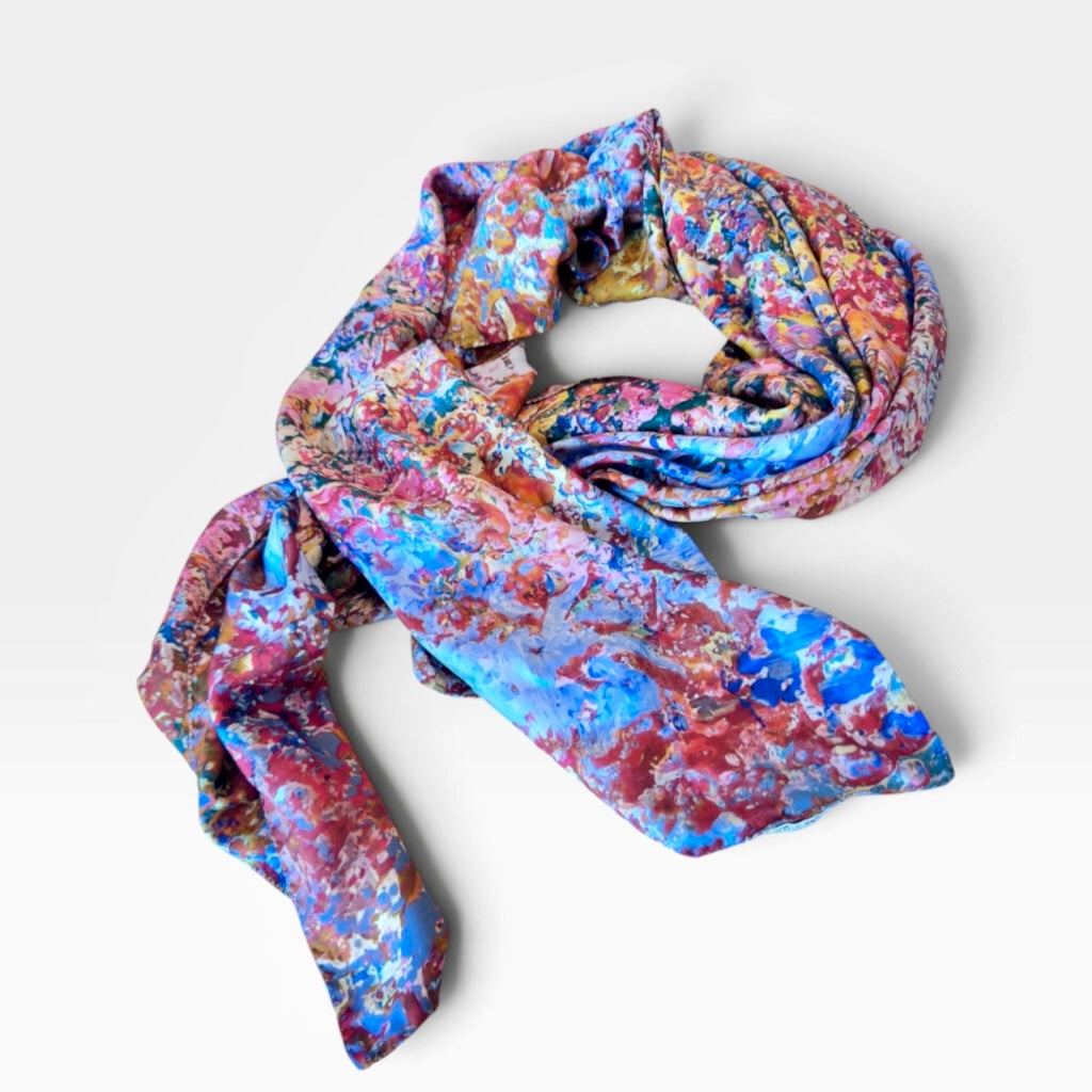 A lightweight scarf with a vibrant, abstract painterly pattern in shades of blue, pink, red, and gold, draped on a light gray background.
